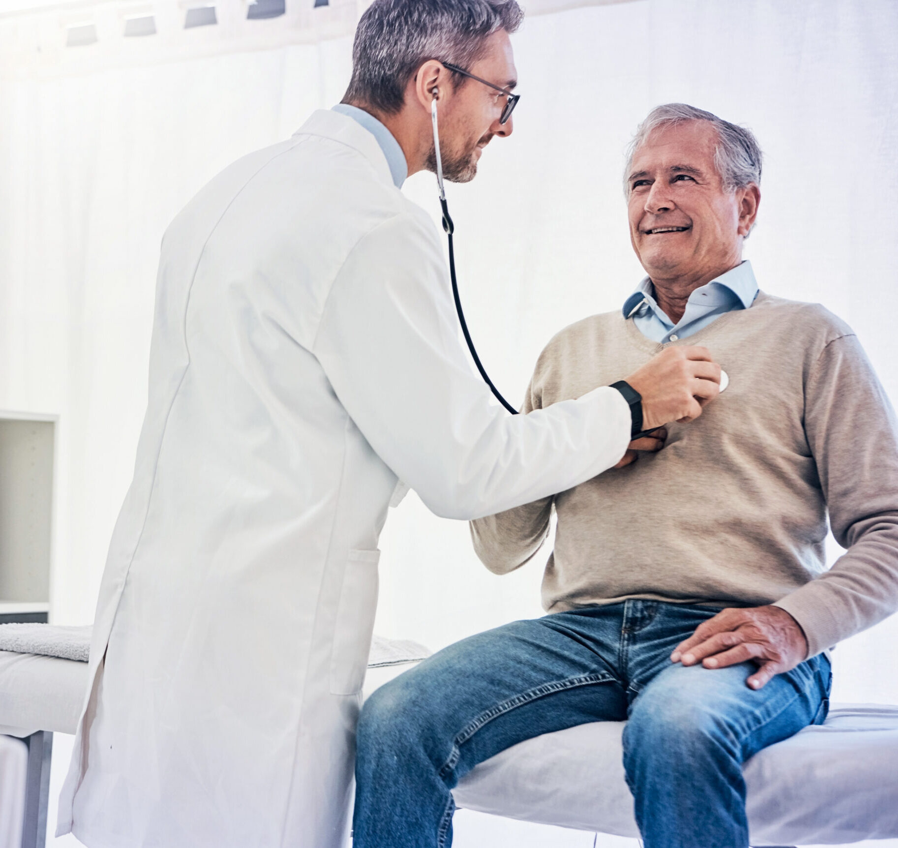 Ein älterer Mann sitzt im Krankenhaus auf dem Bett, während ein Arzt sein Herz abhört.
