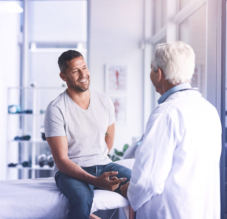Ein junger Mann sitzt beim Arzt auf einer Liege und lässt sich zu Vorsorgeuntersuchungen beraten.