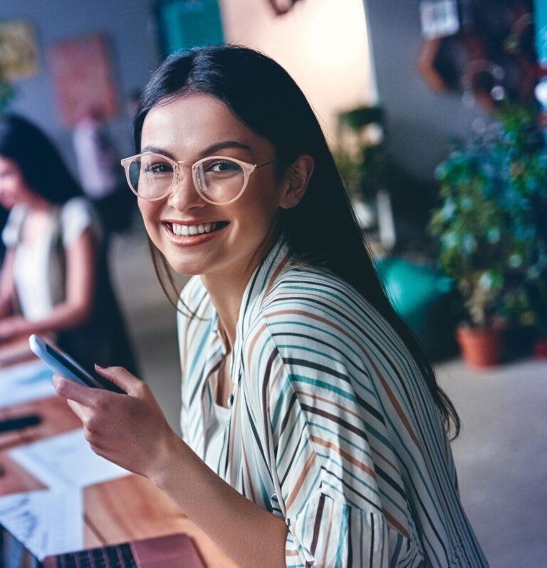 Eine junge Frau sitzt im Büro vor ihrem Laptop und schaut lächelnd in die Kamera.