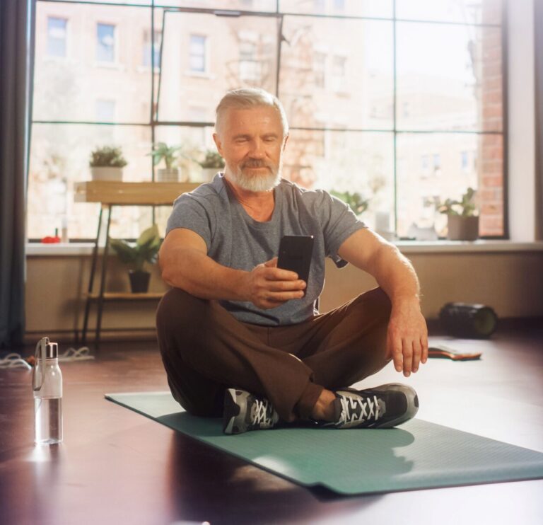 Ein Mann sitzt im Schneidersitz auf einer Yoga-Matte und benutzt eine Gesundheits-App auf seinem Smartphone.