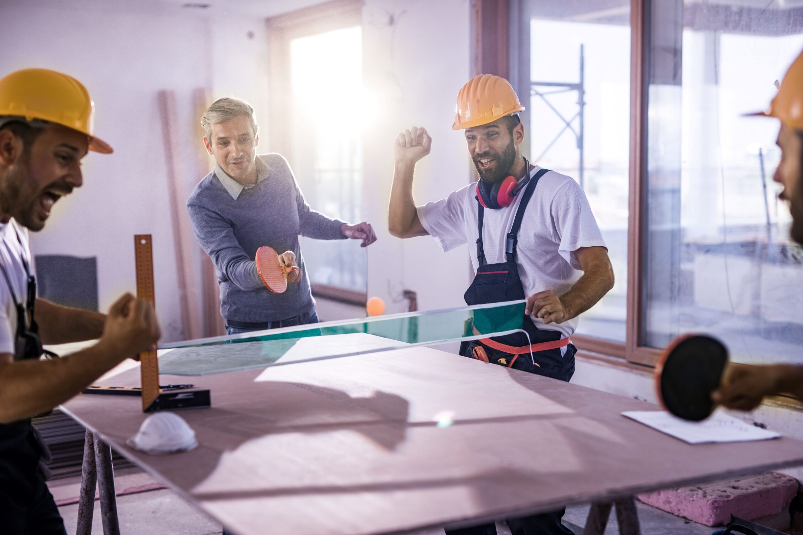 Mitarbeiter mit Bauhelmen spielen Tischtennis auf einer Baustelle.