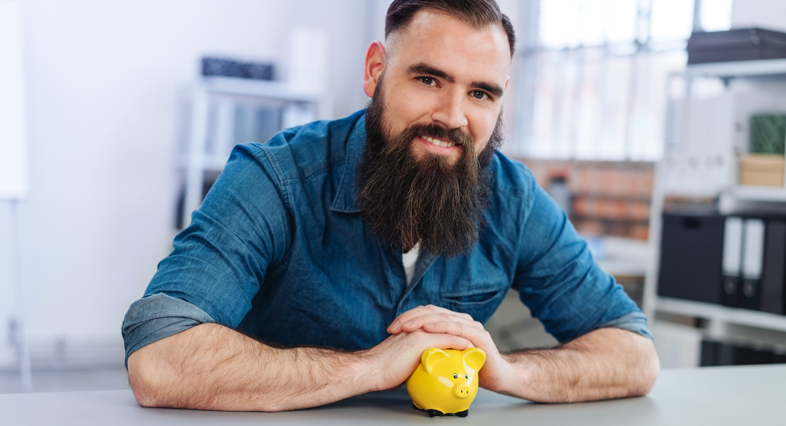 Ein Mann lächelt in die Kamera und hat seine Hände über ein kleines gelbes Sparschwein gefaltet.