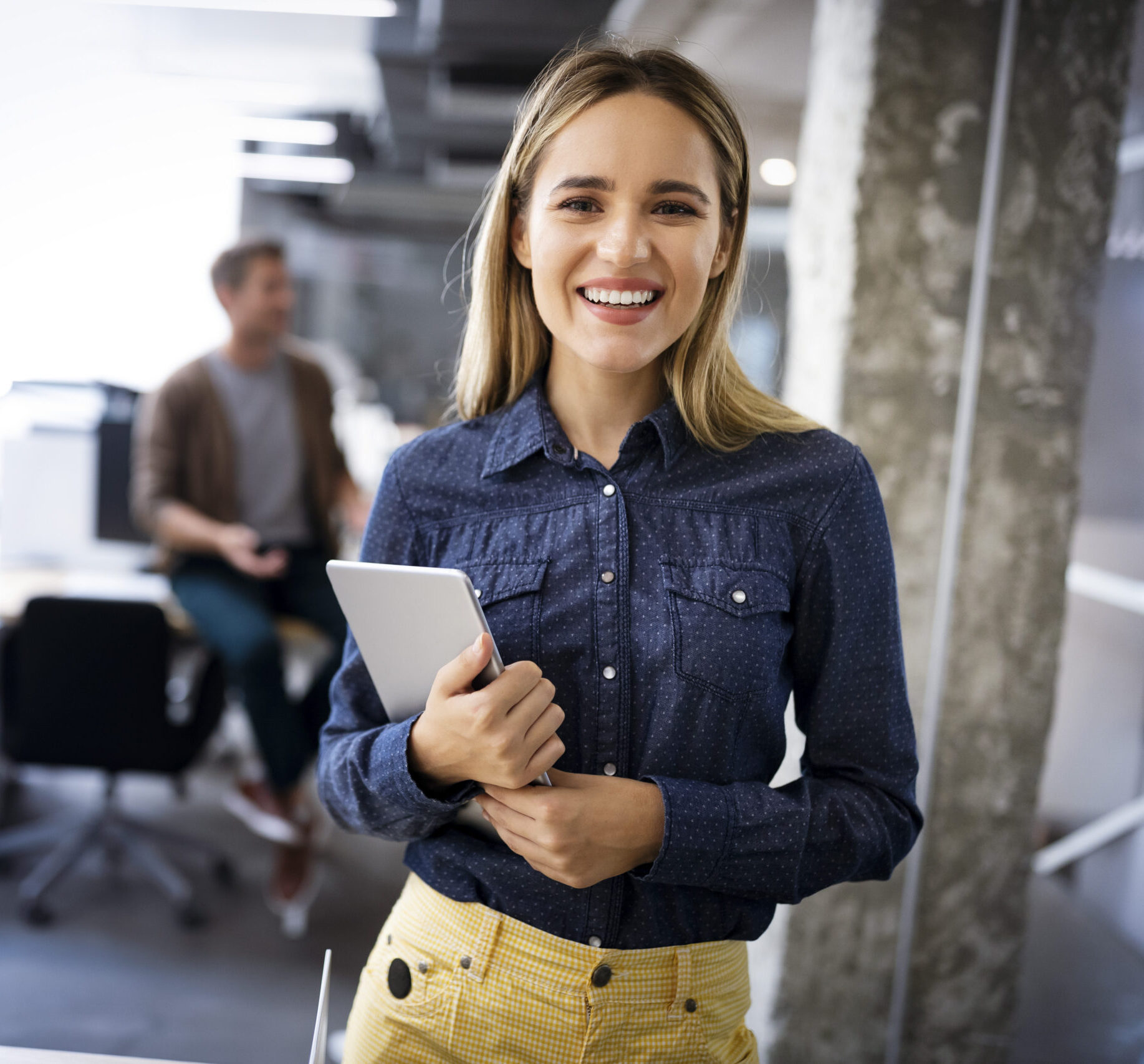Eine junge Mitarbeiterin steht mit Tablet im modernen Büro und schaut in die Kamera.