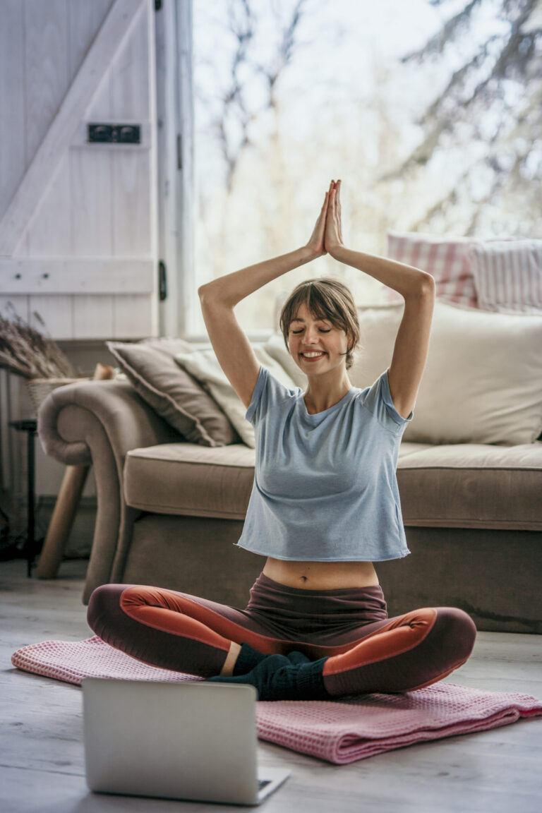 Eine junge Frau folgt einem Yoga-Livestream am Laptop und sitzt in Yogapose auf ihrer Matte im Wohnzimmer.