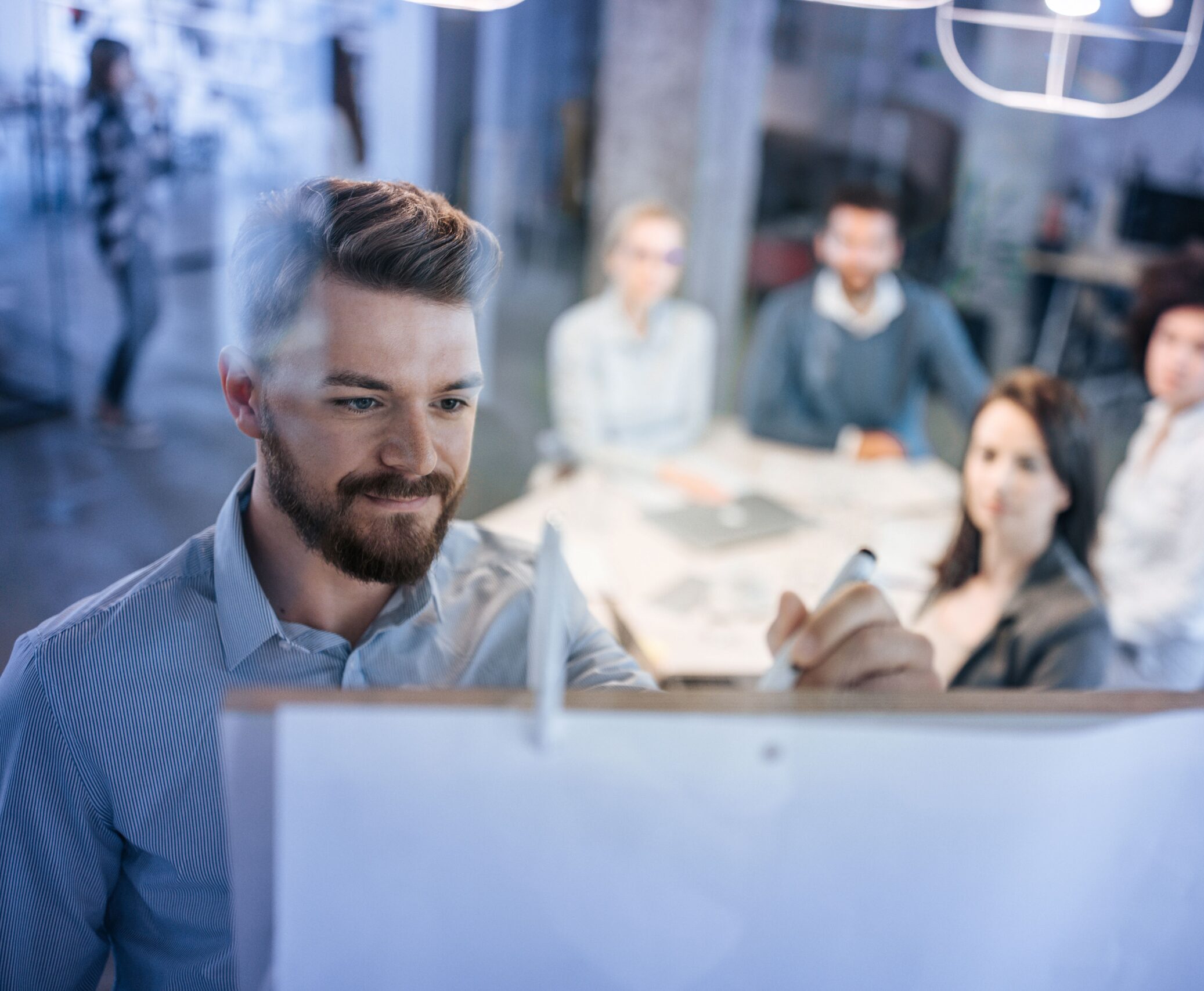 Ein Mann schreibt an ein Whiteboard, während ein Team im Hintergrund aufmerksam zuhört.