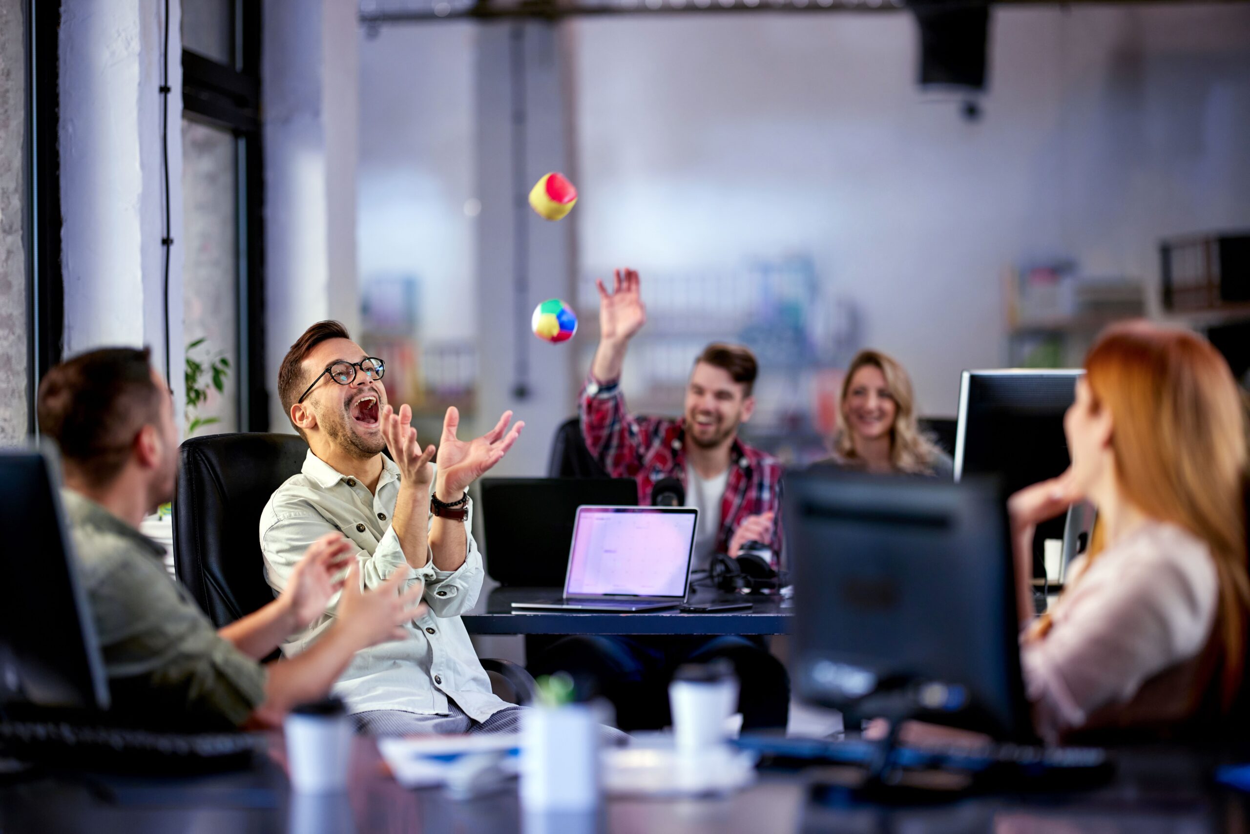 Ein Team im Büro lacht gemeinsam, während ein Kollege mit bunten Bällen jongliert.